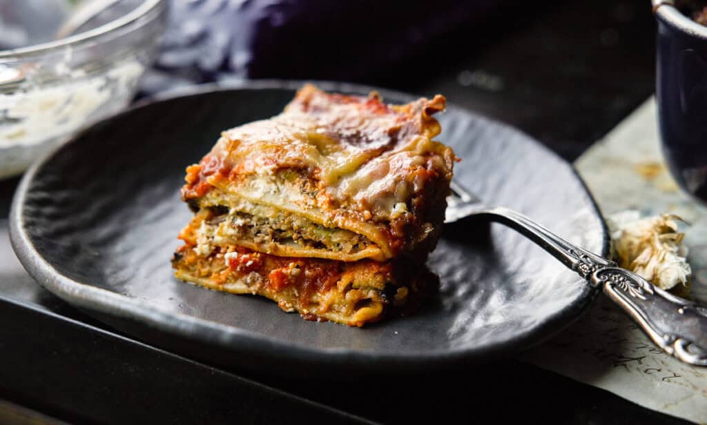 A close-up of a piece of vegan lasagna on a dark plate, with layers of pasta, rich tomato sauce, and melted cheese. A fork rests beside it. The setting has a rustic and cozy feel.